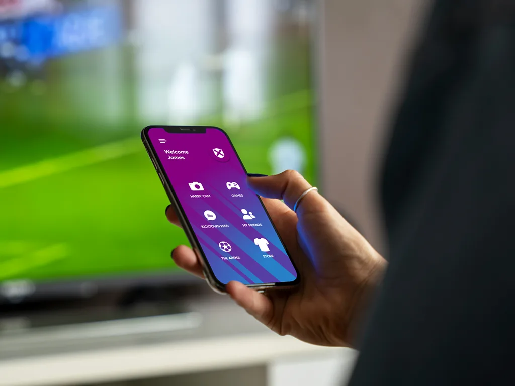 Person holding a phone showing the KickTown app home screen, with the football arena visible behind them